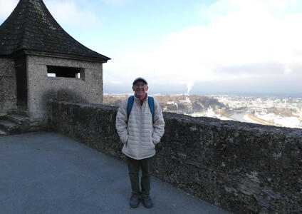 Man with glasses and English driver’s cap, wearing puffy white coat. He is standing at the top of the fortress. At left, a small tower; at right, the city and river below are visible.