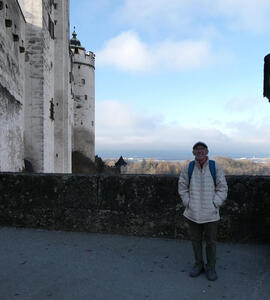 Man with glasses and English driver’s cap, wearing puffy white jacket. To left, a round tower of the fortress.