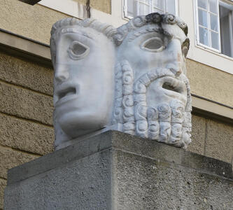 Stone sculpture of heads at right angles. One is a tragedy mask, the other a neutral, bearded figure