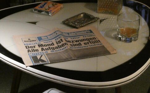 Detail of diorama of people watching moon landing. Newspaper headline: Der Mond is bezwungen: Alle Aufgaben sind erfüllt. A package of Manner Schnitten is at the top of a table, an ashtray in the middle, and a glass of liquor at the right