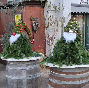Two gnome figures made from evergreen tree branches. They are wearing Santa hats with lights in them and have Christmas ornament balls for noses