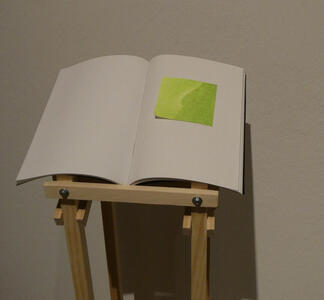 A book, open, on a small stand. The left page is blank; the right page is blank, with a large bright green square “sticky note”.