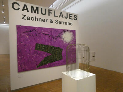Entrance to display; large poster: Camuflajes Zechner & Serrano; in front of poster is an empty birdcage with the door open.