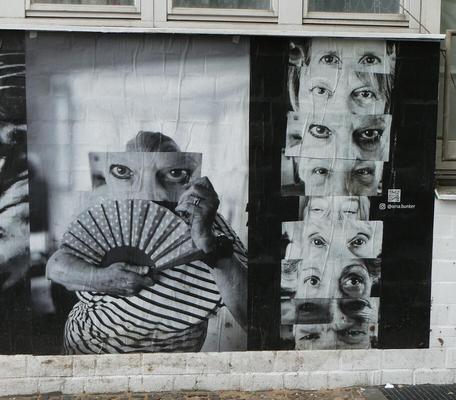 Artist holding a fan in one hand and a large photo of a pair of eyes in front of their face. At right, a vertical set of photos of peoples’ eyes.