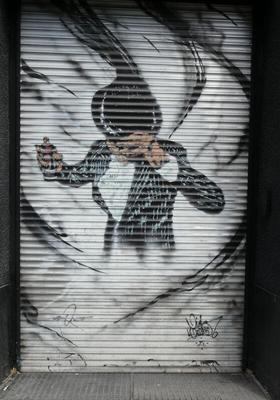 Man in black jacket with white pinstripes, tipping a fedora forwards. In his right hand, a can of spray paint.