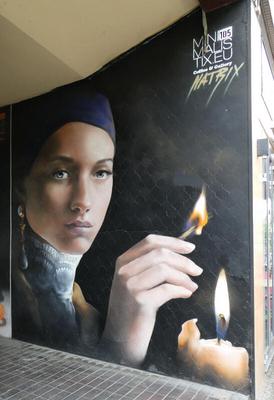 Lady holding a match, lighting a candle. Lady has a bue headscarf and high-necked collar; she is looking at the viewer.