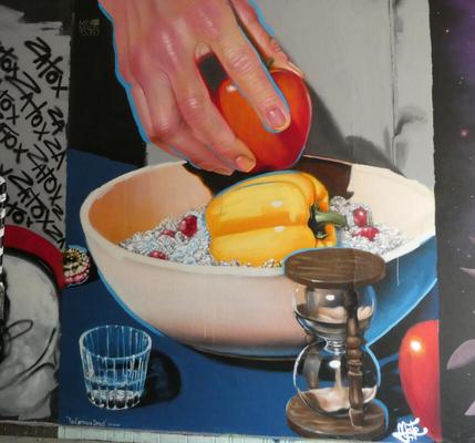 Hand putting an apple into a bowl. A yellow pepper is already in the bowl. In foreground, an hourglass.