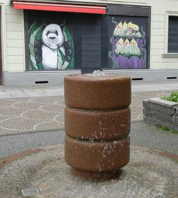 In foreground, a brown cylindrical fountain. In background, a painted giant panda eating leaves.