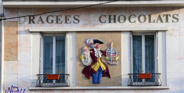 Foppishly dressed man holding baby in one hand and packages of chocolate in other hand.