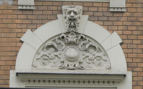 Ornate semi-circular reliefwork with geometric motif at bottom. On top a face with a beard.