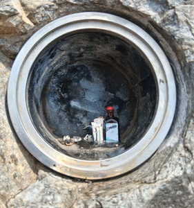 Metal cylinder inside stone wall. Inside the cylinder, a tiny bottle of Cointreau, what look like small pieces of paper, and a couple of pair of googly eyes.