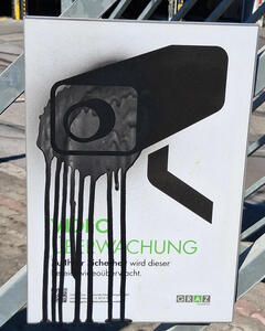Sign saying Video Überwachung with drawing of a video camera. Someone has sprayed black spray paint over the drawing of the camera.
