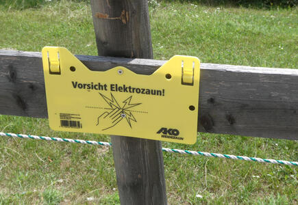 Warning sign: Vorsicht Elektrozaun! with line drawing of hand touching a dotted line and Z-shaped lightning flashes near the fingertips.