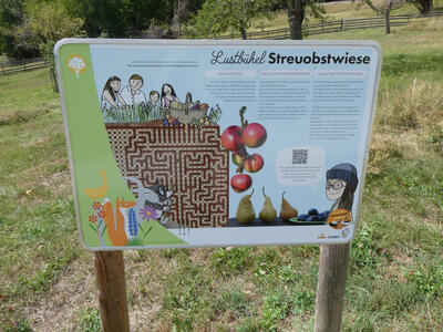 Informational sign about a meadow orchard, oriented towards informing children. There is a maze drawn at the left. People in cartoon style are drawn on the sign.