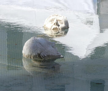 Sculpture of two heads with distressed expressions; they appear to be drowning with their mouths barely above the water.