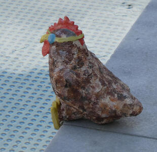 Rooster with stone body, yellow beak, and red comb, sitting at edge of pool