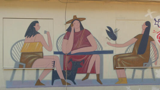 Three women seated around a table. One is drinking a cup of liquid, one is on a phone, and the other has a bird on her arm.
