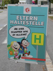 Two smiling children in front of a sign with large letter H in green on yellow. Text: Eltern Haltestelle. One chid is saying Ab hier schaffen wir es alleine! In small print at bottom: Sicher unterwegs. In der kleinsten Großstadt der Welt.