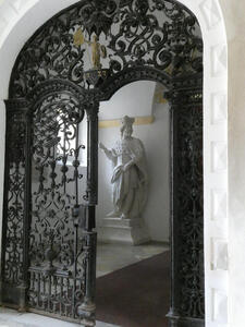 Arch with wrought iron gate. In background, a statue of a holy person.