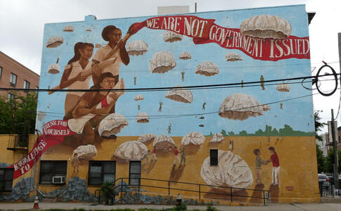 Young women, one holding diploma, another a paintbrush with a strip of paint reading “We are not government issue”.  Multiple parachutes in background with text such as Our humanity, our futures, our dreams