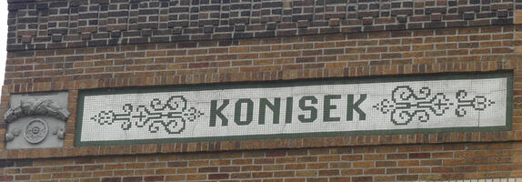 Name “Konisek” spelled out in black mosaic tiles with white tile background on a brick building.