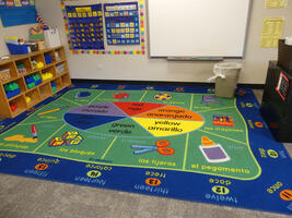 Floor rug labeled with colors, numbers, and classroom supplies; labels in Spanish and English
