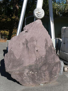 Large brown stone. Atop it, a round stone carving of a face with a toothy grin.