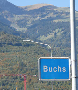 Highway sign, blue with white lettering: Buchs. Mountain in background.