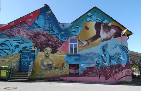 Painted building wall. At left, a woman with flowing hair At right, a man being squeezed in by a window frame and the top of the frame he is in.