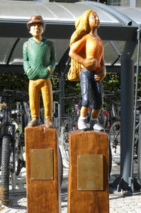 Lifeike wood statues of a man in green sweatshirt and long-haired woman in yellow shirt and blue jeans