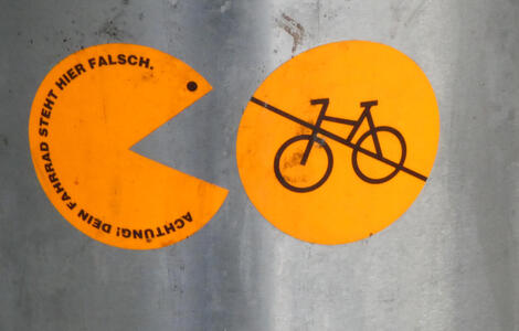 Sticker at left is a Pac-Man style character. Tet: Achtung! Dein Fahrrad steht hier falsch. To its right, a circular sticker with a picture of a bicycle and a diagonal slash through it.