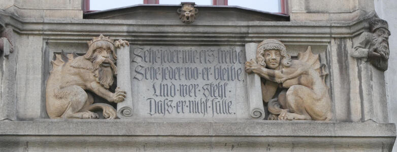 Two stone dragons with human faces (one male, one female), holding a scroll with German words on it.