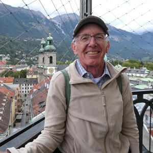 Man in glasses with English driver’s cap. In background, church and mountains.