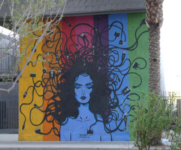 Woman with blue face on rainbow background; her hair ends in computer connector cables.